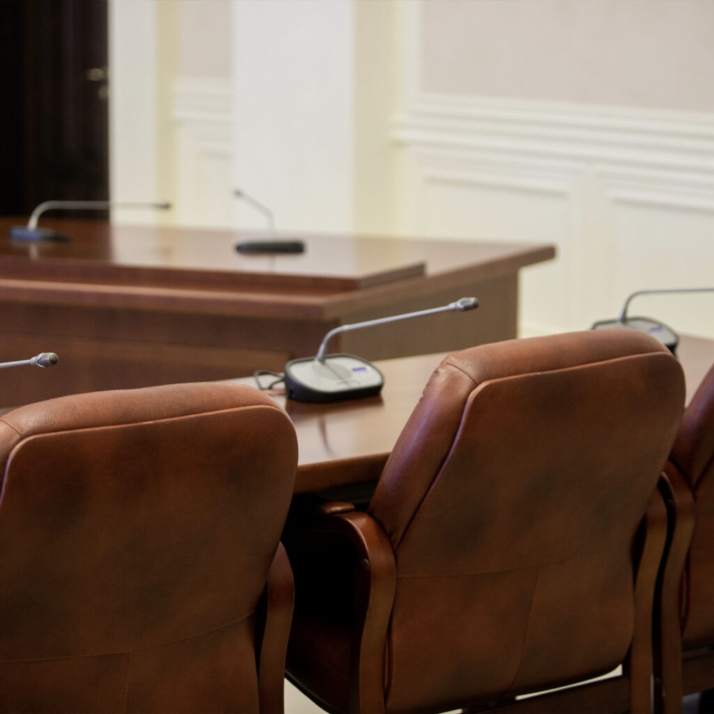 Brown leather chairs and wooden desks with microphones arranged in a conference or meeting room setting, with a neutral-colored wall and molding in the background.