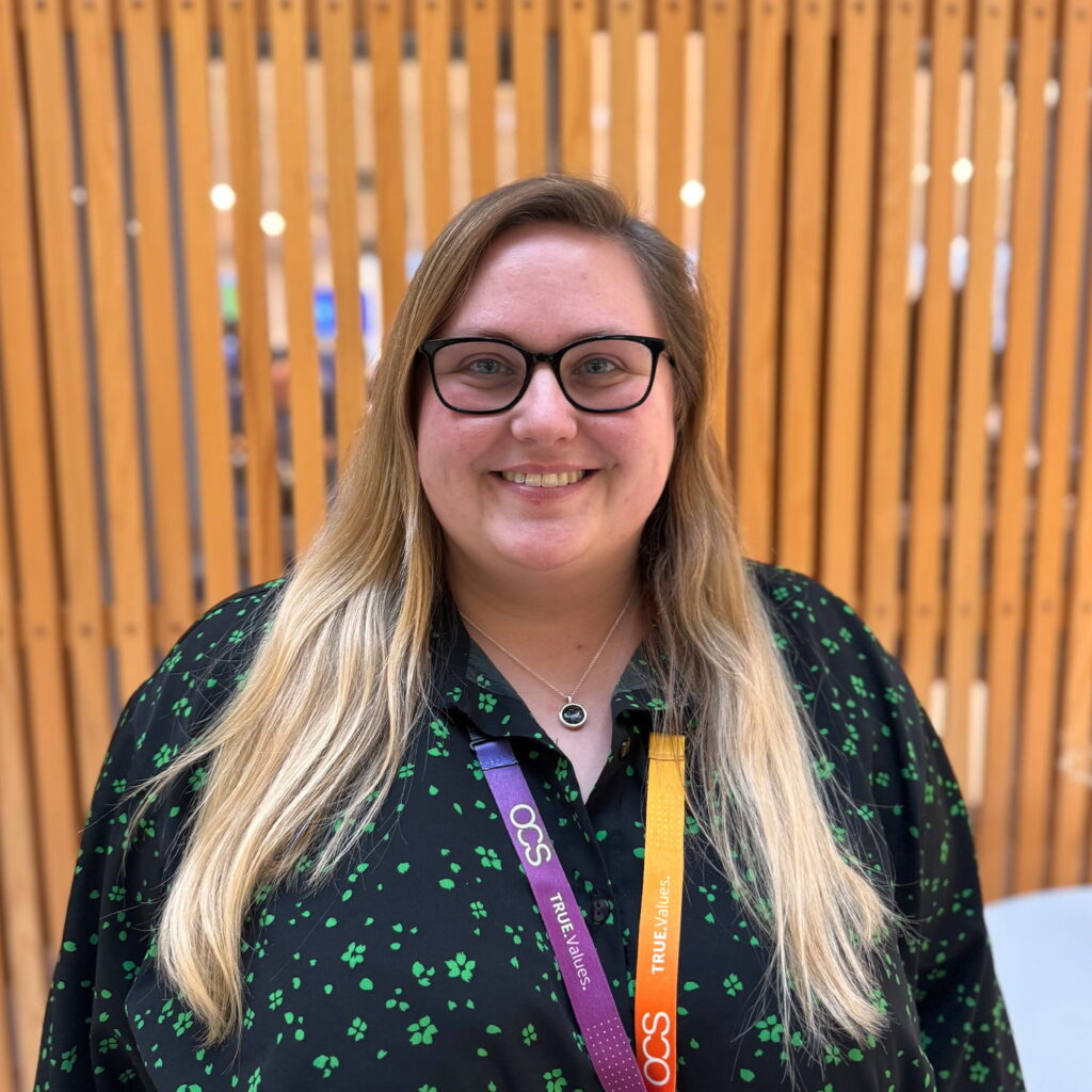 A person with long blonde hair, glasses, and a black top with green patterns stands smiling in front of a wooden slat wall, wearing colorful lanyards around their neck.