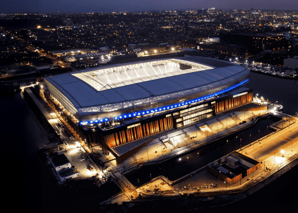 Aerial view of a brightly lit stadium at night, surrounded by water and city lights, with seating areas illuminated and empty seats visible inside.