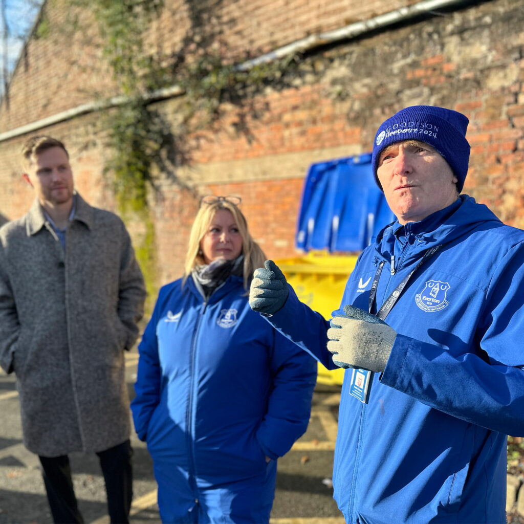 Three people stand outdoors near a brick wall and yellow bin; two in blue jackets and hats, one in a gray coat. The man in front gestures with his hands, while sunlight casts strong shadows.