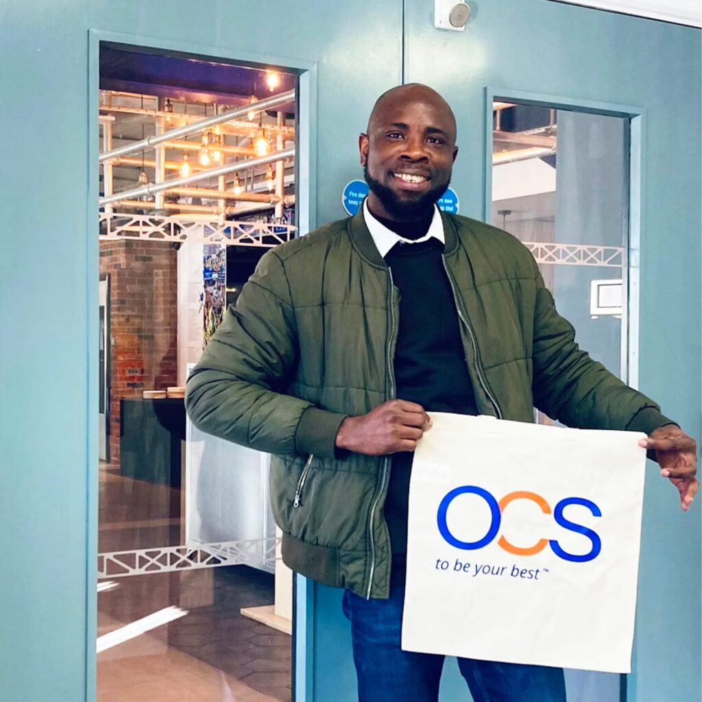 A smiling man in a green jacket stands indoors, holding a white OCS bag with the slogan to be your best™ in blue and orange letters. Glass doors and a modern interior are visible behind him.