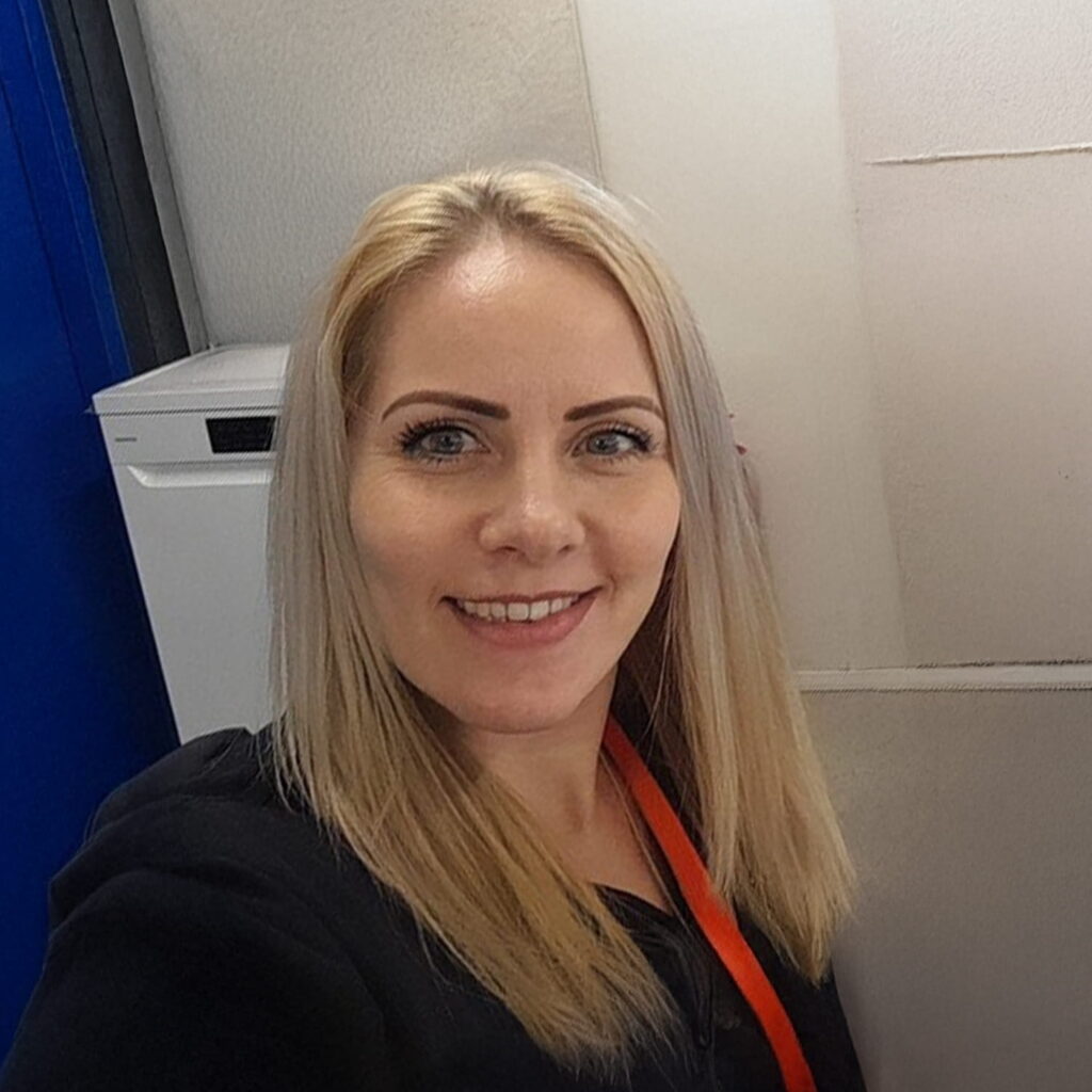 A woman with straight blonde hair, light skin, and blue eyes is smiling at the camera. She is wearing a black top and a red lanyard, standing indoors against a white and blue background.