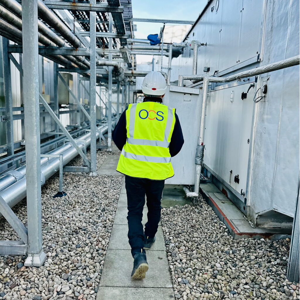A person wearing a high-visibility vest labeled OCS and a white hard hat walks along a gravel path between industrial buildings and metal pipes.