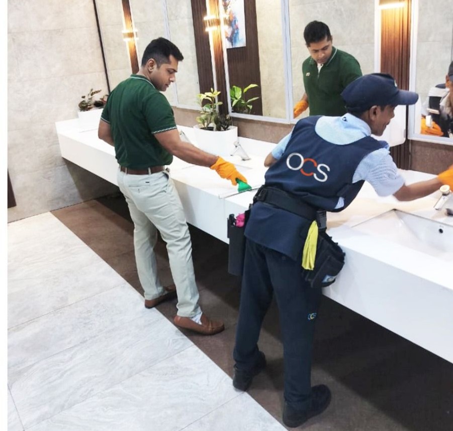 Two men clean a public restroom sink area; both wear green shirts and orange gloves, one has cleaning supplies attached to a vest. A large mirror and plants are visible on the countertop.