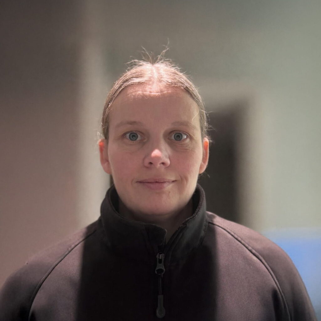 A person with light skin and straight, light brown hair tied back, wearing a black zip-up jacket, stands indoors against a blurred background. They are looking directly at the camera with a neutral expression.