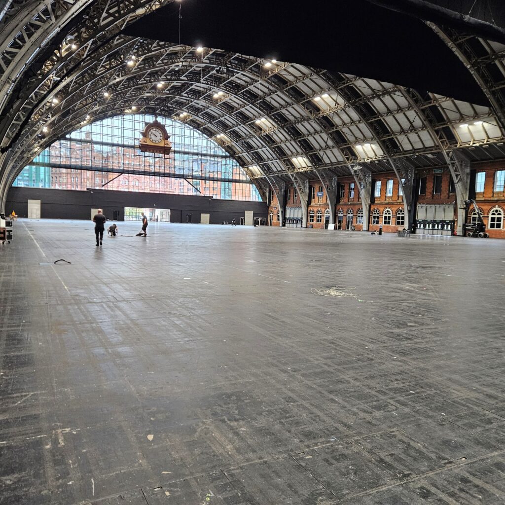 A large, empty indoor hall with a high arched ceiling, exposed beams, and a big clock above a glass wall at the far end. A few people are walking across the spacious floor.