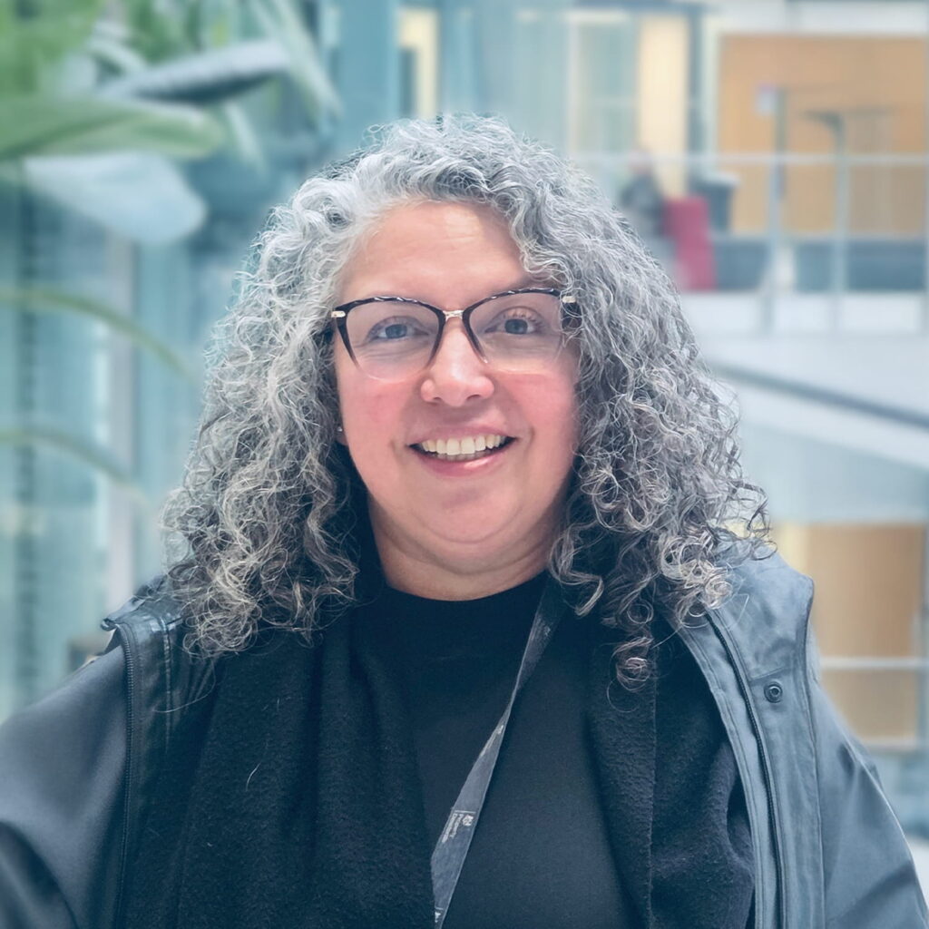 A woman with curly gray hair and glasses smiles at the camera. She is wearing a dark top and jacket, standing indoors with blurred office and plant background.