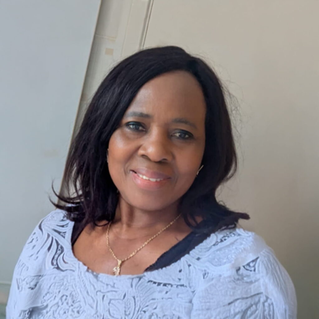 A woman with shoulder-length dark hair smiles at the camera. She is wearing a light purple patterned top and a gold necklace. The background is a light-colored wall.