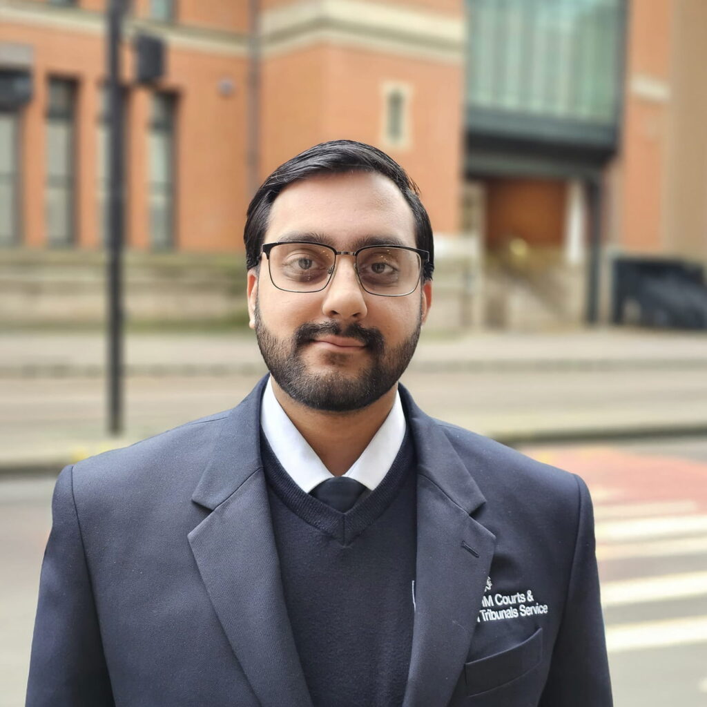 A man with dark hair, a beard, and glasses, wearing a suit jacket and sweater, stands outside on a city street in front of a brick building.