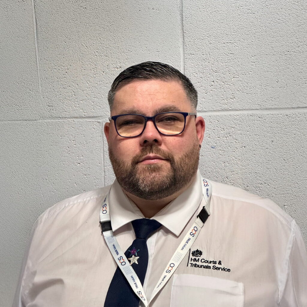 A man with short hair, a beard, and glasses wears a white shirt, dark tie, and HM Courts & Tribunals Service lanyard, standing against a light grey textured wall.