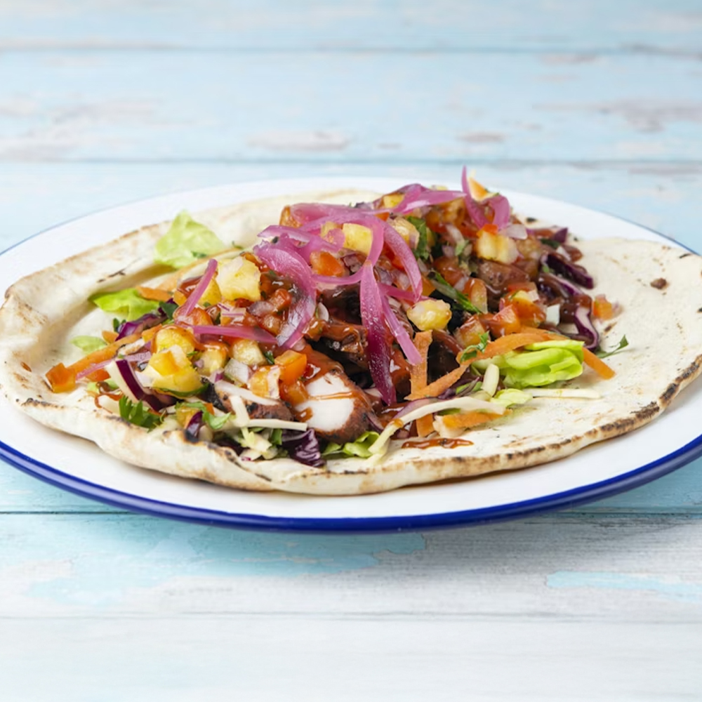 A flatbread topped with grilled chicken, shredded lettuce, diced tomatoes, pineapple salsa, red onions, and sauce, served on a white plate with a blue rim on a light blue wooden table.