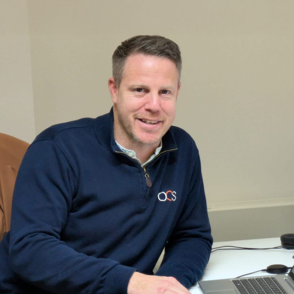 A man with short hair and a beard is sitting at a desk, smiling at the camera. He is wearing a navy blue sweater with a small embroidered logo and has a laptop and computer mouse in front of him.