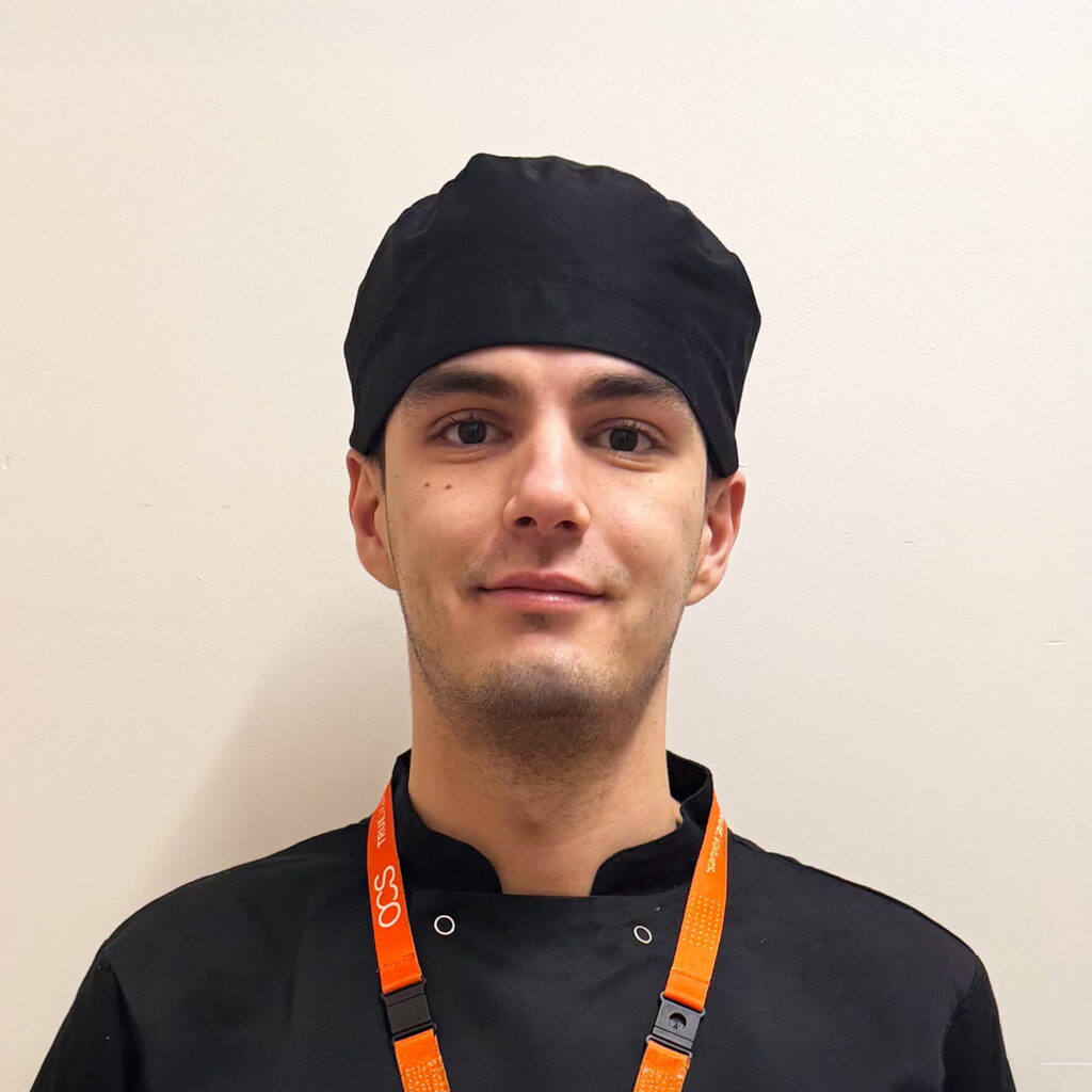 A young person wearing a black chefs hat and jacket with an orange lanyard stands against a plain light-colored wall, looking at the camera with a neutral expression.