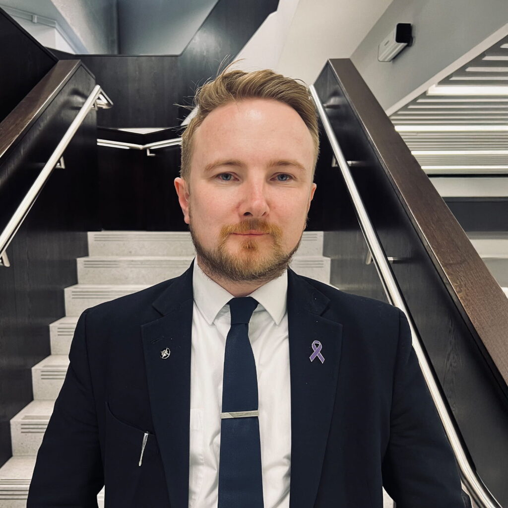 A man with blonde hair and a beard, wearing a black suit, white shirt, and navy tie, stands in front of a modern staircase. He has a purple awareness ribbon pinned to his lapel.