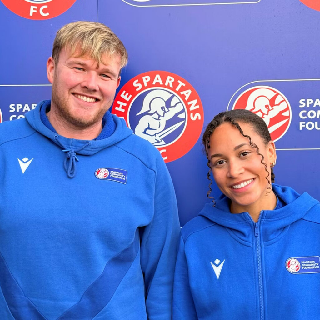 Two people wearing blue Spartans Community Foundation hoodies stand smiling in front of a blue wall with Spartans FC and foundation logos.