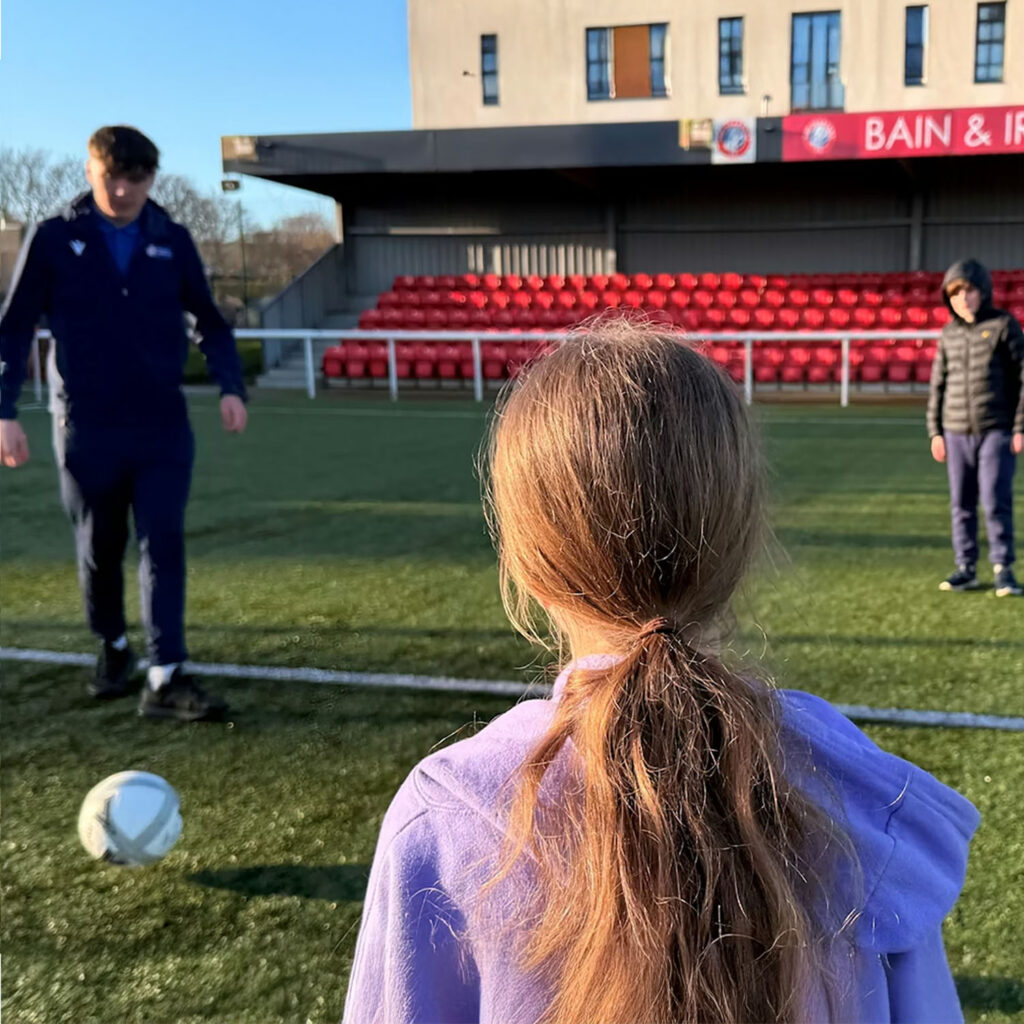 A child with long hair in a ponytail stands on a soccer field, watching a man in sportswear approach a soccer ball, while another child stands nearby. Red stadium seats and a building are in the background.