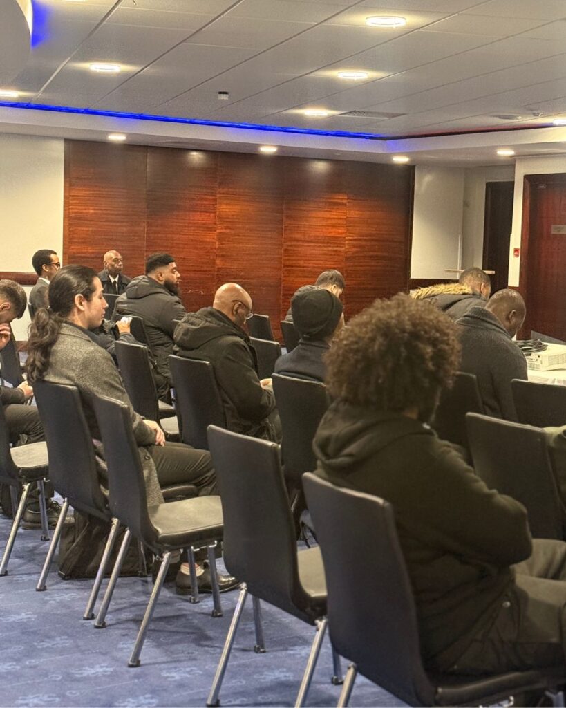 A group of people sit spaced out in rows of black chairs in a conference room with wood-paneled walls and blue carpeting, listening attentively to an unseen speaker.