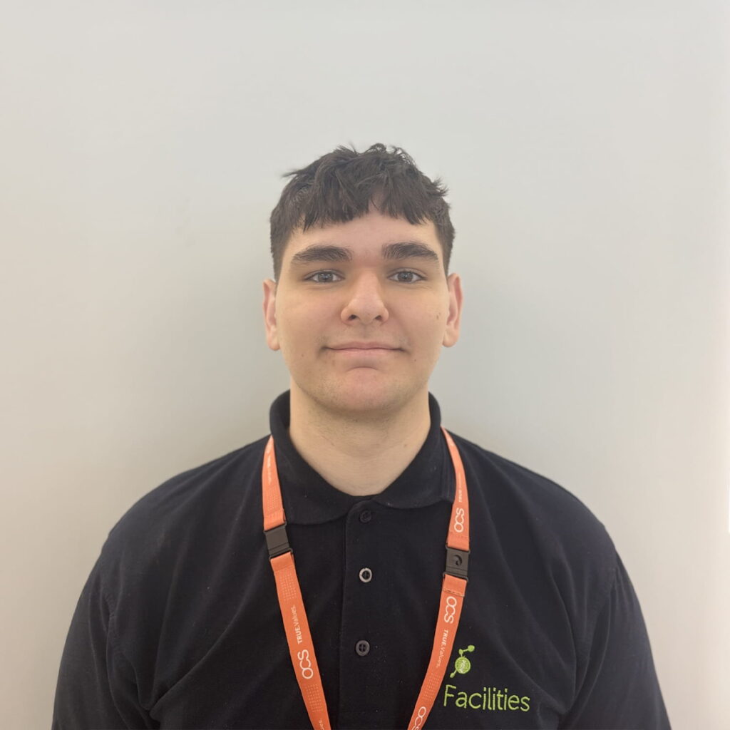 A young man with short dark hair wears a black polo shirt with a Facilities logo and an orange lanyard. He stands against a plain white background, looking straight at the camera with a neutral expression.