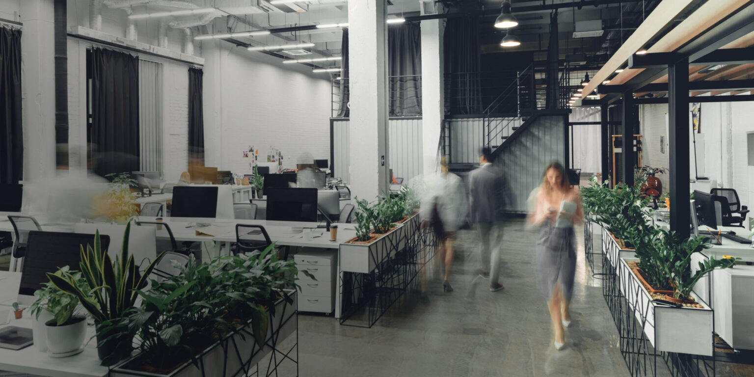 A modern open-plan office with several desks, computers, and plants. Some people are walking through the space, creating a motion blur effect. The office is bright with high ceilings and industrial-style decor.