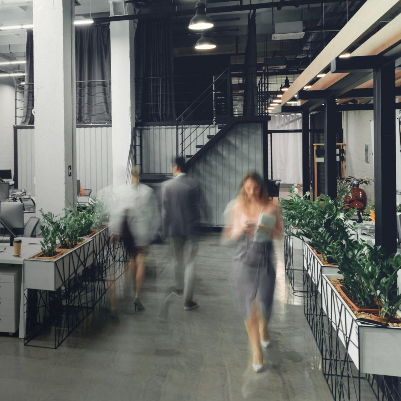 Modern open-plan office interior with blurred figures of people walking, rows of desks with computers and green plants, and a black staircase leading to an upper level in the background.