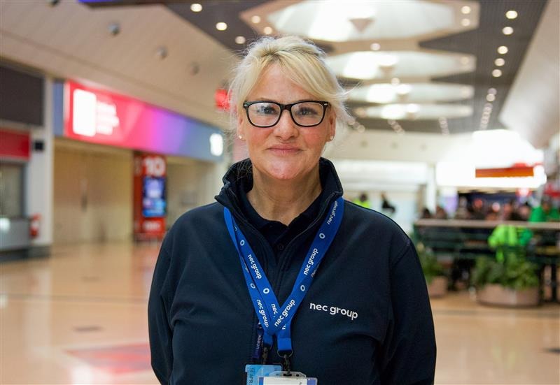 A woman with blonde hair, glasses, and a blue NEC Group sweatshirt stands indoors. She wears a blue lanyard with ID badges around her neck, smiling slightly in a well-lit corridor.