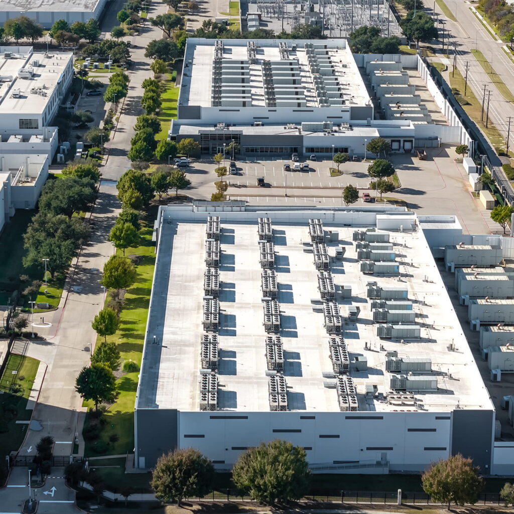 Aerial view of a large industrial facility with white rooftops featuring rows of HVAC units, surrounded by green lawns, trees, roads, and nearby buildings.