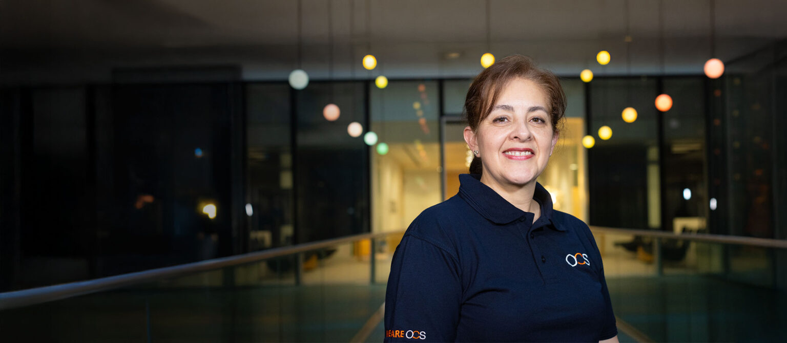 A woman with light brown hair and a dark blue polo shirt stands smiling in a modern, dimly lit interior with colourful hanging lights in the background.