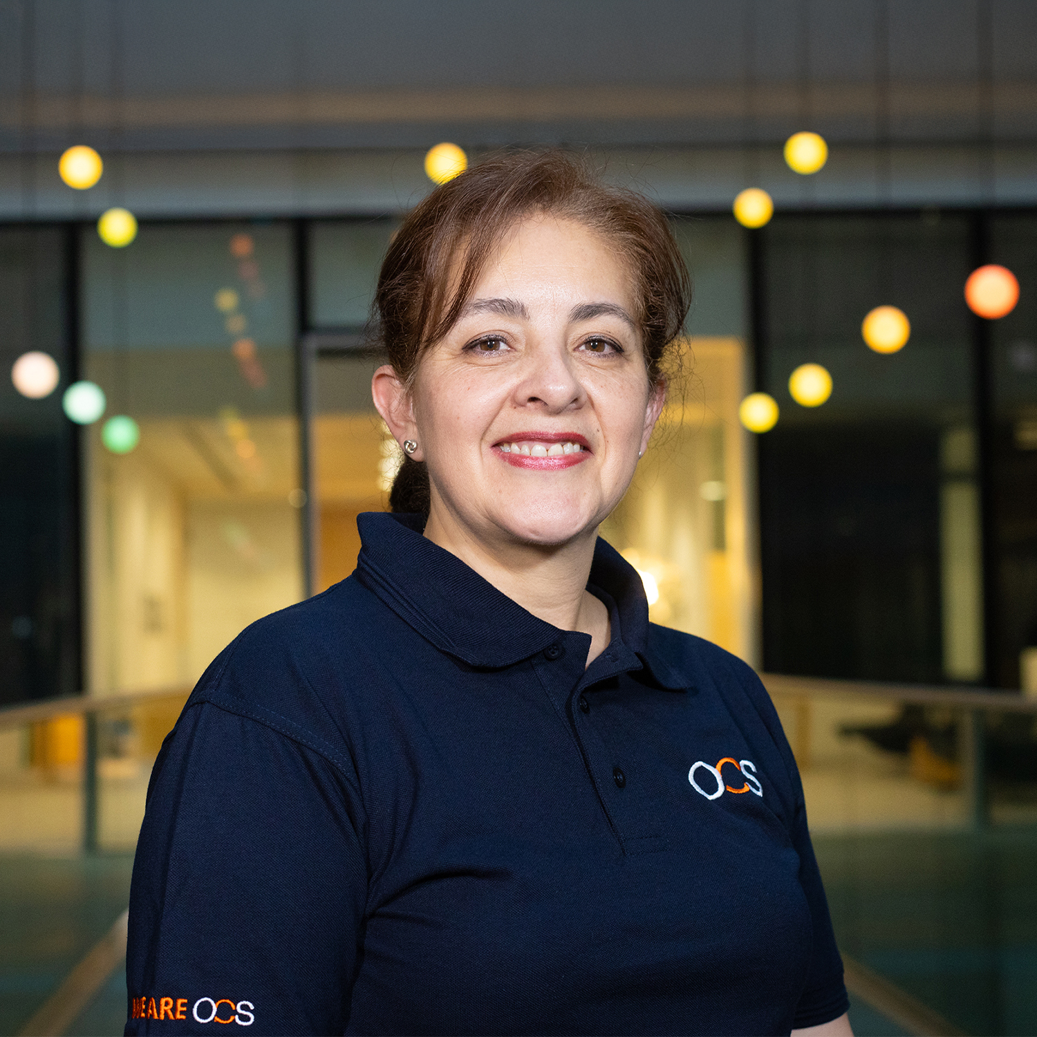 A woman with light brown hair, wearing a navy blue OCS polo shirt, smiles at the camera. She stands indoors in front of large glass windows with hanging colourful lights in the background.
