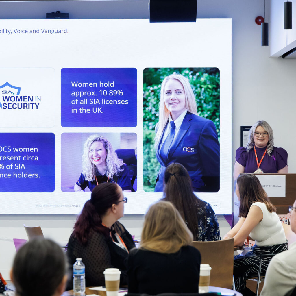 A speaker presents to an audience of women at a conference. Behind her, a screen displays information and photos about women in security, including statistics on women holding SIA licences in the UK.