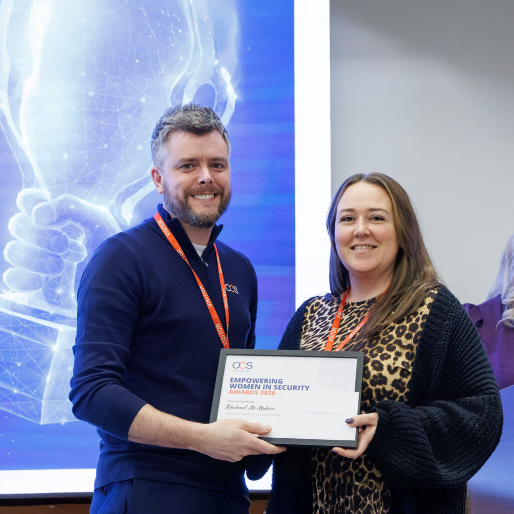 A man and a woman smiling and holding an Empowering Women in Security Award certificate. Both wear conference lanyards and stand in front of a digital, blue-lit background.
