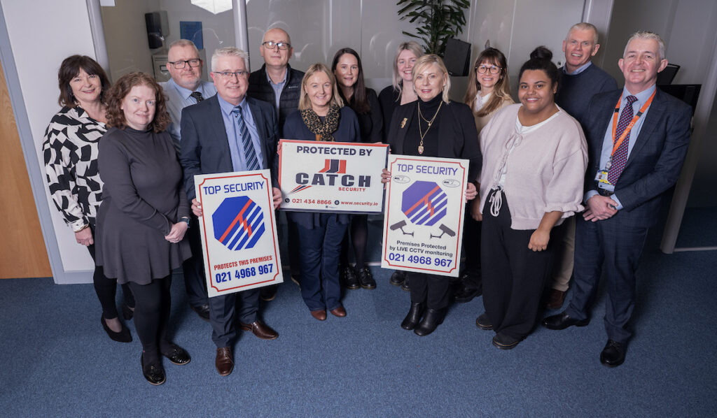 A group of thirteen people stand indoors, smiling at the camera. Three people in front hold security signs that read “PROTECTED BY CATCH” and “TOP SECURITY,” with contact information displayed.