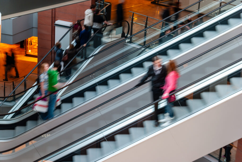 People using escalators in a busy indoor shopping centre. Some individuals appear blurred, indicating movement, while others stand still. Glass railings and modern architecture are visible in the background.