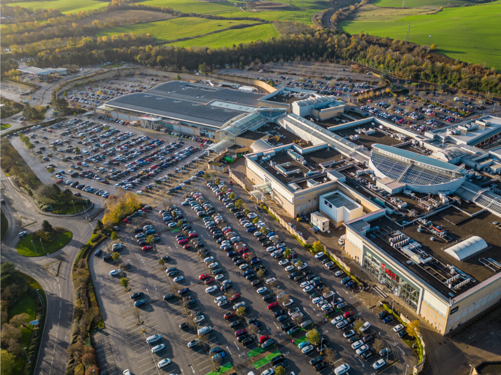 Aerial view of a large shopping centre with a vast car park filled with cars, surrounded by green fields and trees on a sunny day.