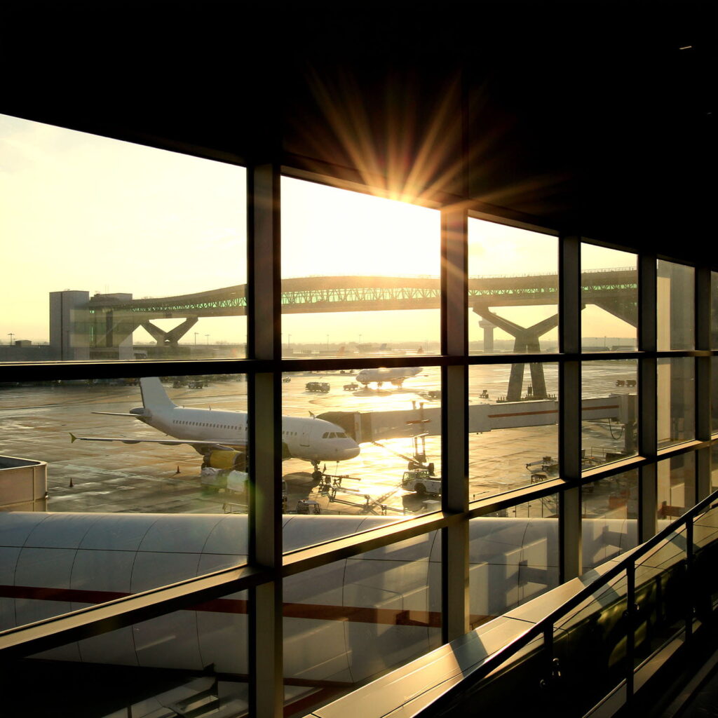 Sunlight streams through large airport windows, illuminating an aeroplane parked at the gate on the tarmac. The scene captures the early morning or late afternoon light, with a bridge visible in the background.