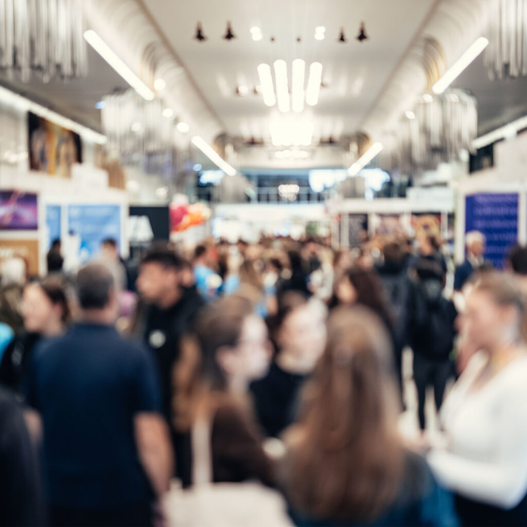 A large group of people gather and socialise in a brightly lit, modern indoor space with banners and stands, suggesting a busy event or exhibition. The image is blurred, making faces and details indistinct.