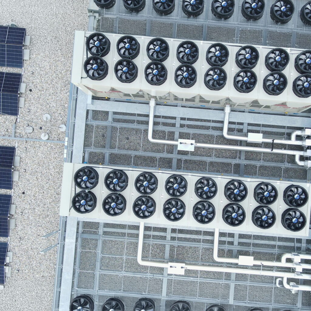 Aerial view of large rooftop cooling units with multiple circular fans and pipes, next to several solar panels, all situated on a gravel-covered building roof.