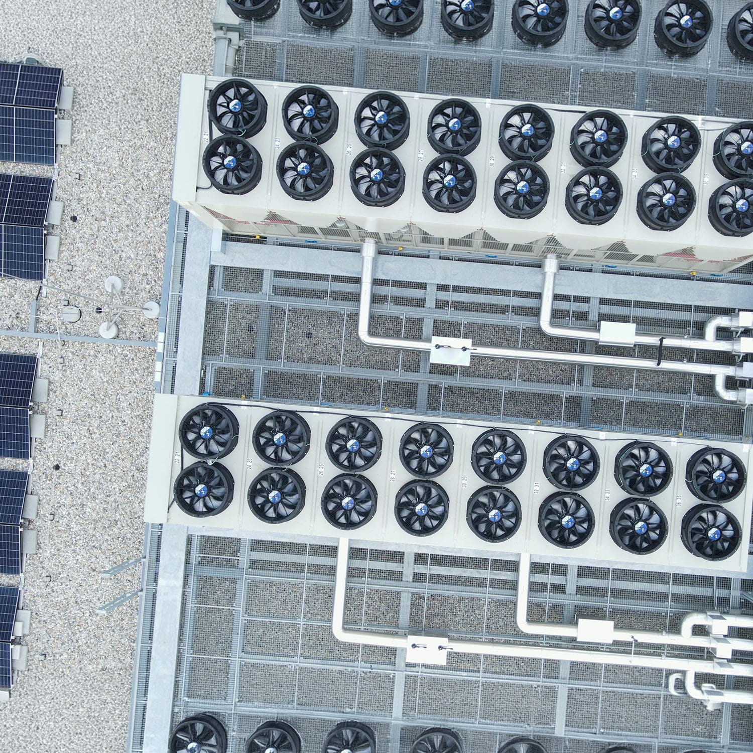 Aerial view of large rooftop cooling units with multiple circular fans and pipes, next to several solar panels, all situated on a gravel-covered building roof.