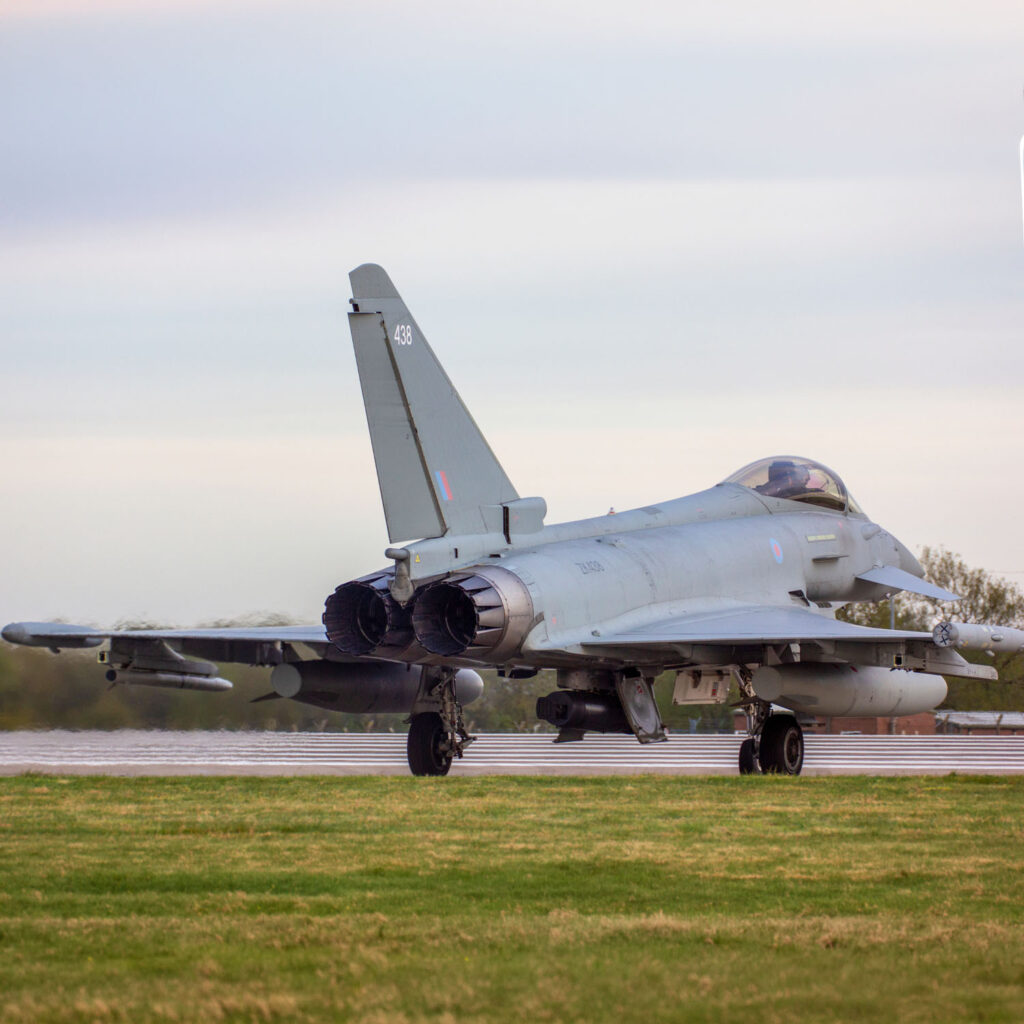 A military fighter jet, identified as a Eurofighter Typhoon, is taxiing on a runway with its rear engines and tail fin visible. The aircraft is grey and set against a cloudy sky and grassy field.