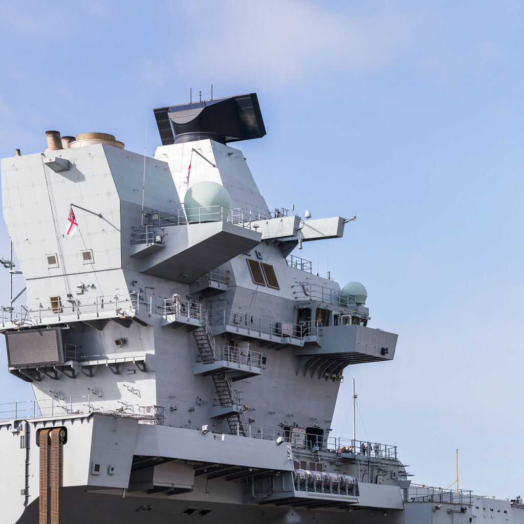 The image shows a close-up view of the superstructure of a modern naval aircraft carrier, featuring large radar domes, aerials, various decks, ladders, and communication equipment against a clear blue sky.