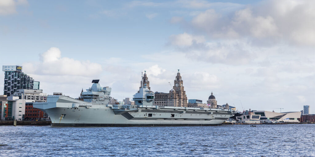 A large grey aircraft carrier is docked by a city waterfront, with modern and historic buildings in the background under a partly cloudy sky. The water in the foreground is calm.