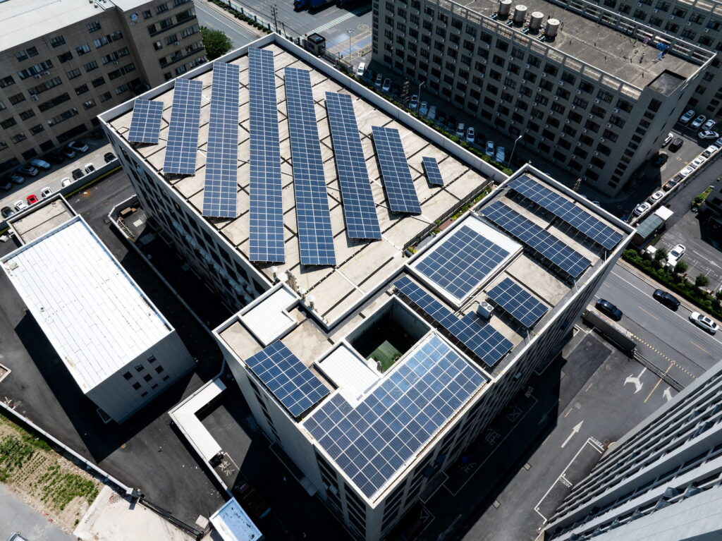 Aerial view of several large buildings with numerous solar panels installed on their rooftops, surrounded by paved roads and parked cars in an urban setting.