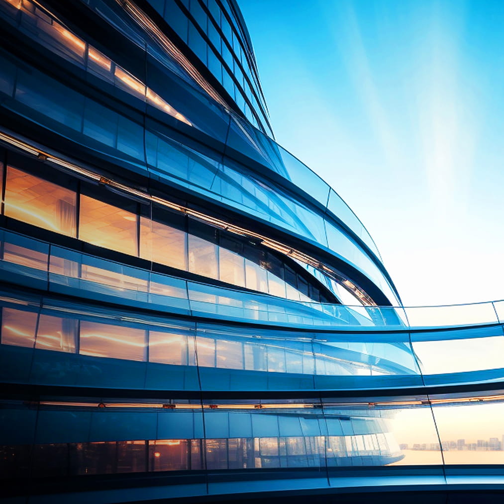 Curved glass facade of a modern building reflecting sunlight, with multiple storeys and a clear blue sky in the background.
