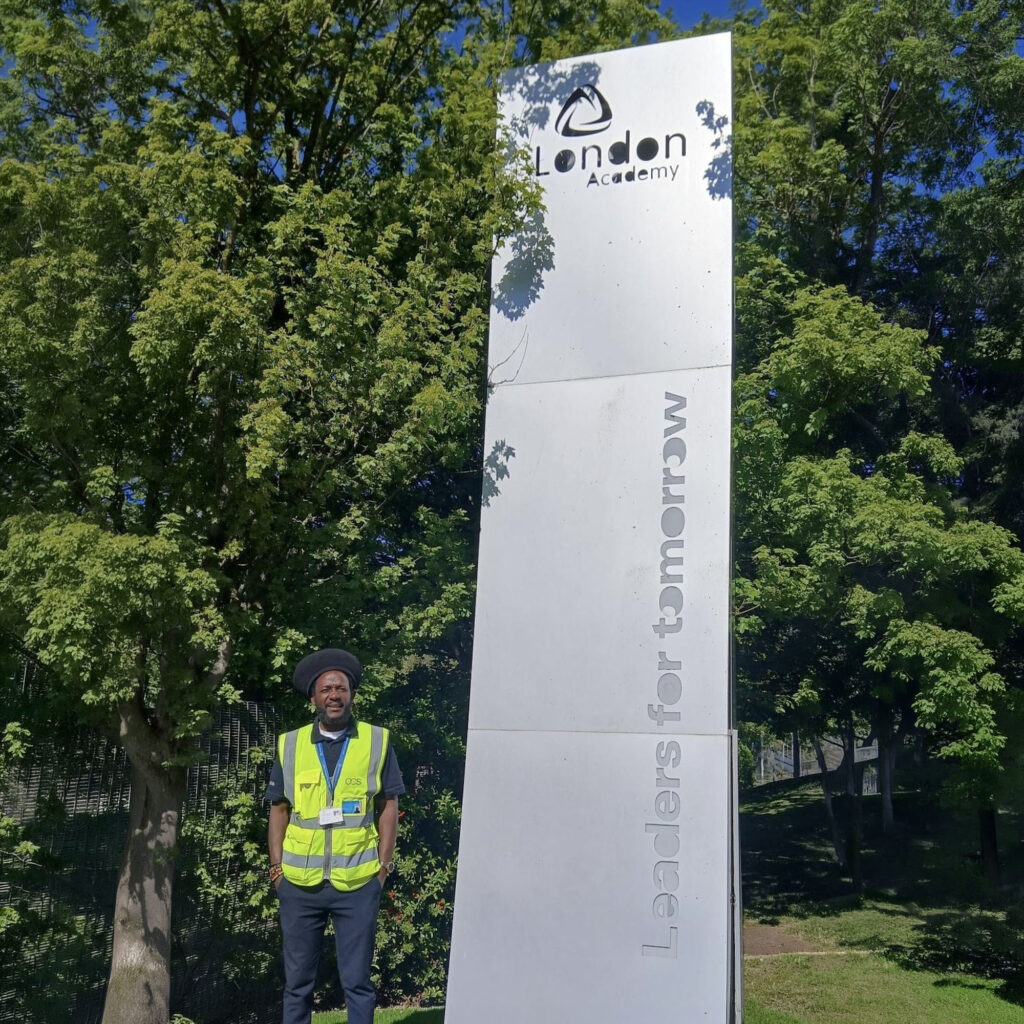 A person wearing a high-visibility vest stands next to a tall silver sign that reads London Academy and Leaders for Tomorrow, with trees and greenery in the background.