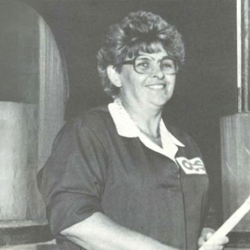 A woman with short curly hair and glasses, wearing a dark collared shirt, stands indoors and smiles. The photo is in black and white.