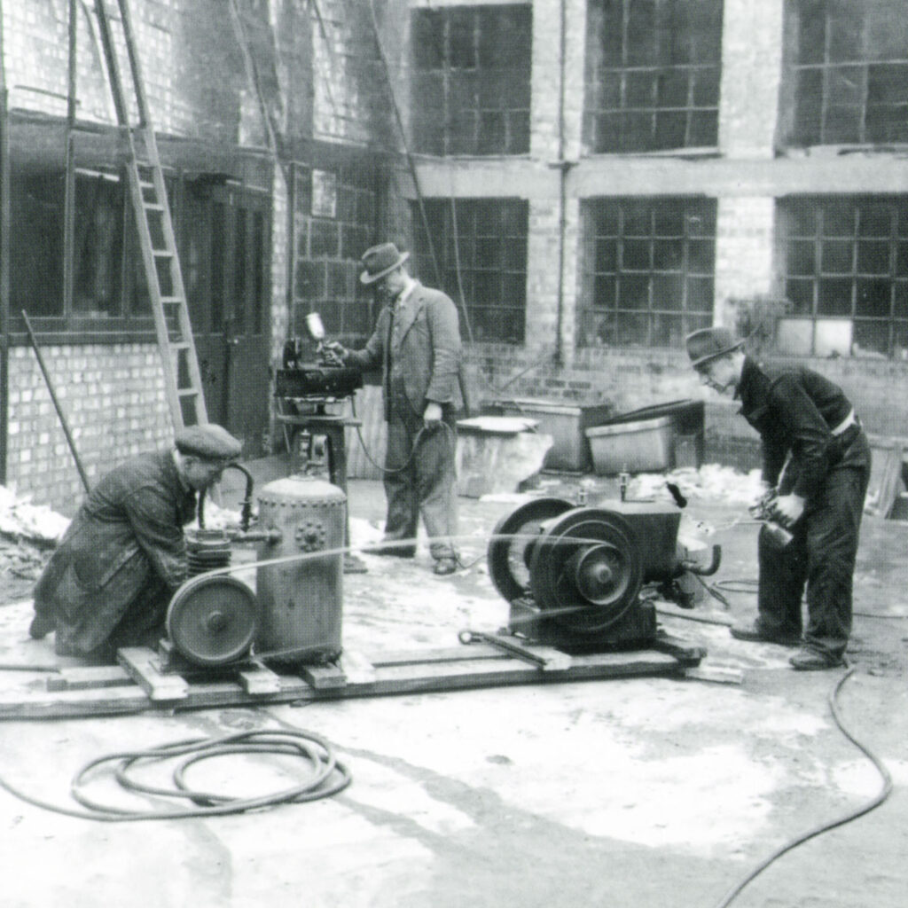 Three men work with early 20th-century industrial machinery in an outdoor courtyard with brick walls and large windows. Two men kneel beside engines while another stands at a machine in the background.