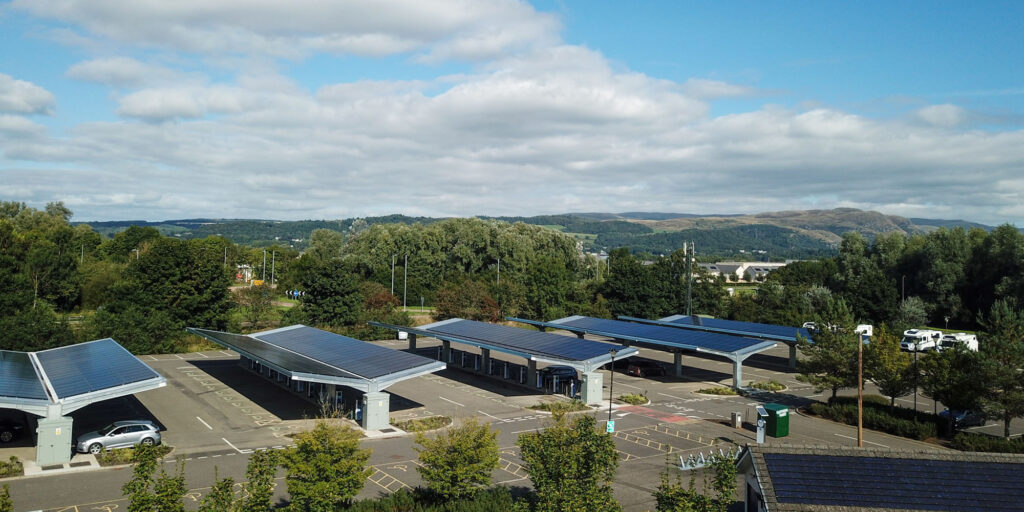 A car park with several carports, each topped with solar panels. Trees surround the car park, and hills and clouds are visible in the background under a blue sky.