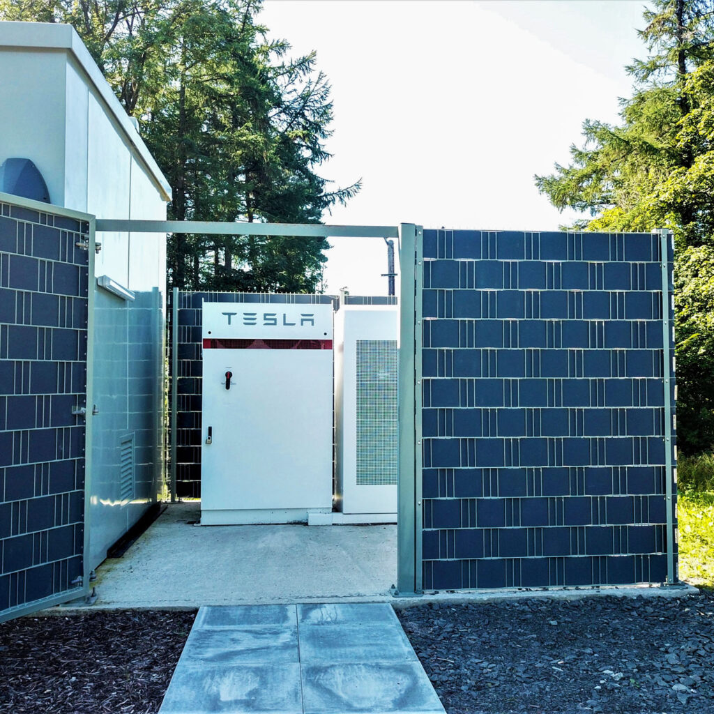 A Tesla energy storage unit is installed outdoors, surrounded by a fence made of solar panels and trees in the background. The unit is placed on a concrete plinth next to a building.