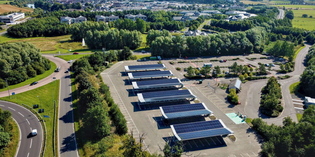 Aerial view of a car park with solar panel canopies over parking spaces, surrounded by trees and roads, with buildings and a town visible in the background.