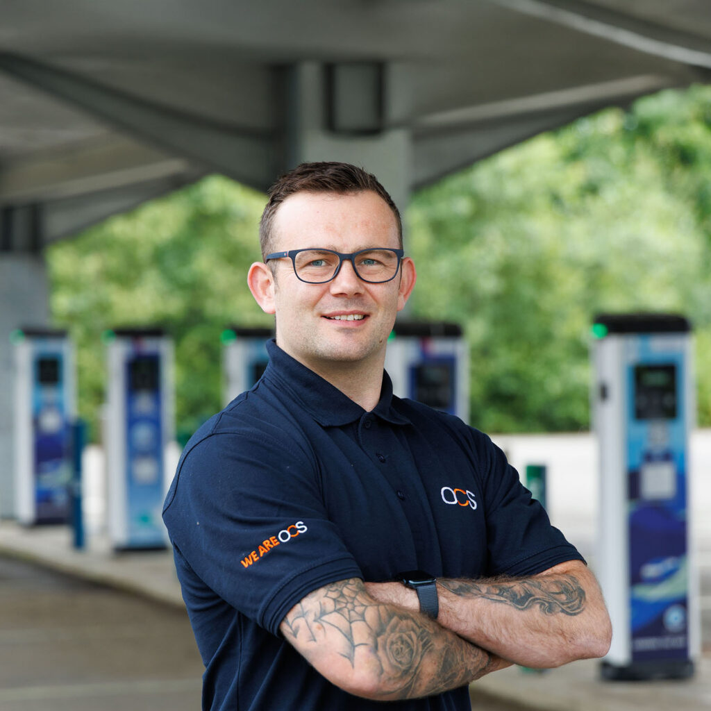 A man with glasses and tattoos, wearing a navy WE ARE OCS polo shirt, stands with arms folded at an outdoor electric vehicle charging station. Blurred green trees and multiple chargers are visible in the background.