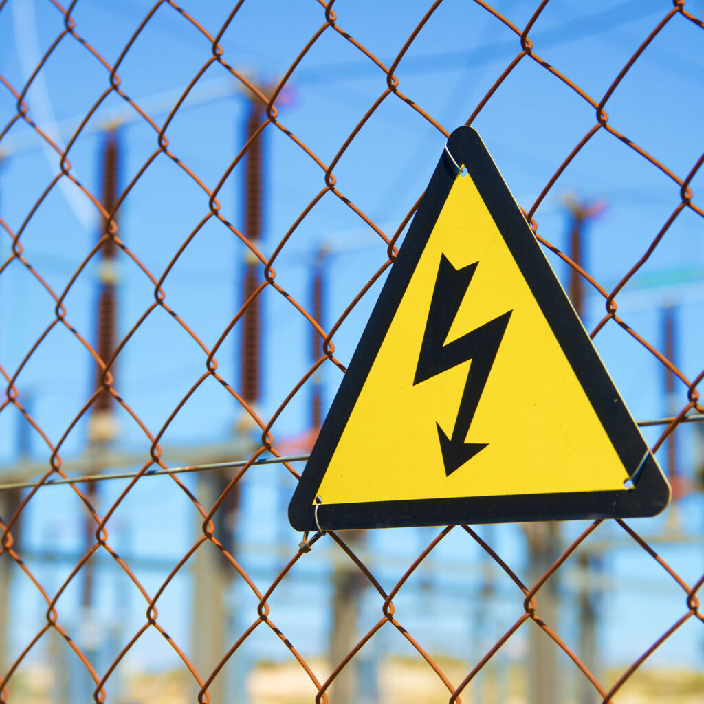 A yellow triangular high voltage warning sign is attached to a rusty wire fence, with electrical substation equipment and a clear blue sky visible in the background.